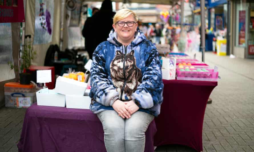 Mujer sentada al borde de su puesto en el mercado, con una colección de cajas detrás de ella, usando una chaqueta con cremallera con la foto de un perro husky