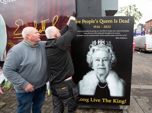Dos hombres con un pequeño mural negro con una foto en blanco y negro de la reina y las palabras 