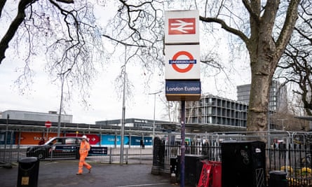 Un hombre con un traje de alta visibilidad camina por el bordillo de una calle muy transitada junto a un cartel que muestra el antiguo símbolo de British Rail y un símbolo de metro