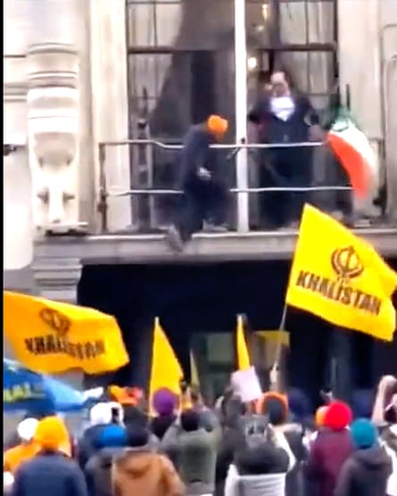 la gente ondea banderas amarillas mientras el hombre se aferra al balcón del primer piso de la Alta Comisión India